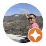 A man in sunglasses and a pink shirt smiles and waves toward the distant Hollywood sign, standing near a railing with a camera resting on it. Hills and clear blue sky are in the background. An orange star badge overlays the bottom right corner. Fuji Omakase Sushi San Fernando Valley